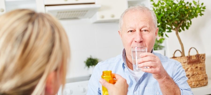 An elderly man sitting in a bright kitchen drinks a glass of water while a caregiver hands him medication from a prescription bottle.