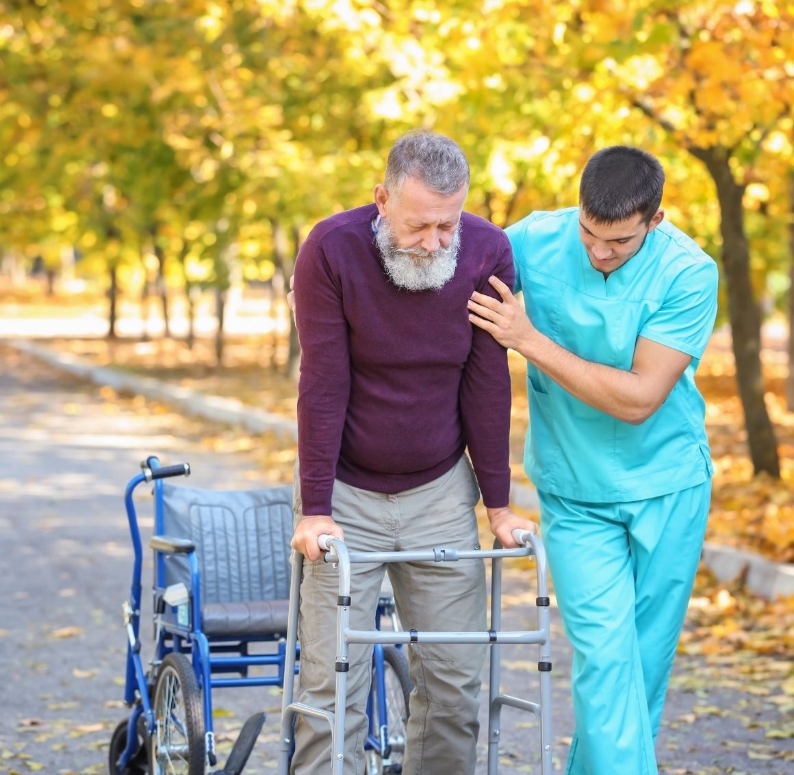 Caregiver assisting a senior man with mobility support during an outdoor walk A caregiver in teal scrubs supports an elderly man using a walker along a tree-lined path. A wheelchair is positioned behind them, emphasizing safe mobility assistance, encouragement, and compassionate outdoor care.
