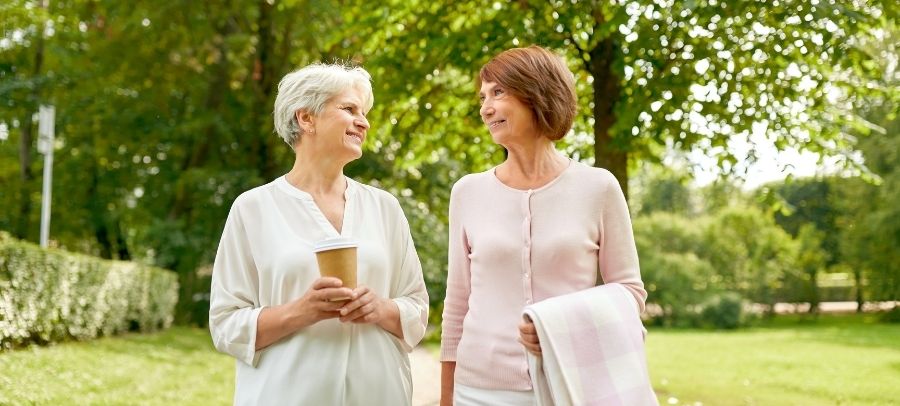 Relaxing Outdoor Stroll and Companionship for Seniors Two older women walking together in a park on a sunny day, smiling and talking. One holds a coffee cup, and the other carries a blanket.