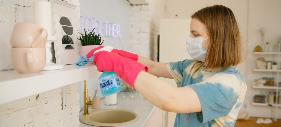 A caregiver wearing a face mask and pink cleaning gloves wipes a shelf while holding a spray bottle in a tidy kitchen. The scene reflects attention to cleanliness, sanitation, and supportive in-home housekeeping.