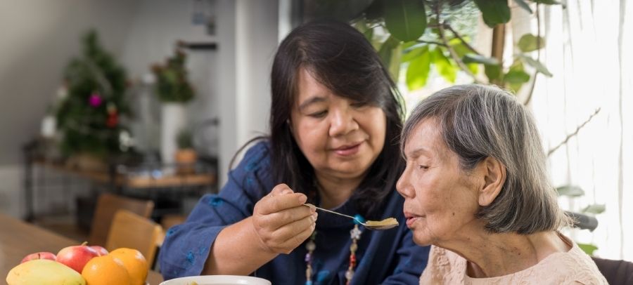 Compassionate Caregiver Assisting Senior Woman with Mealtime Support A caregiver gently feeds an elderly woman at a dining table, offering warm and attentive mealtime assistance in a home setting.