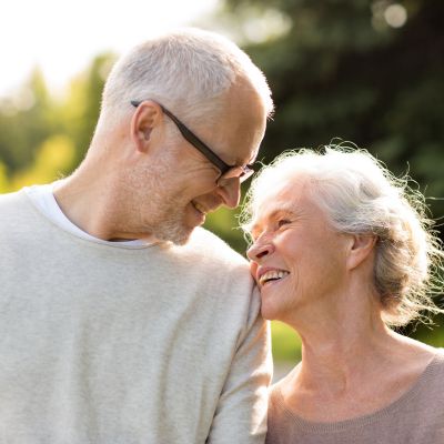 A joyful senior couple standing outdoors, smiling warmly at each other as the woman leans affectionately on the man's shoulder in a sunlit, natural setting.