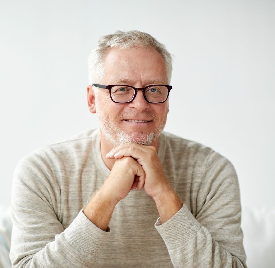 A relaxed, smiling elderly man with glasses and a light sweater rests his hands together while sitting indoors. The image reflects confidence, comfort, and well-being.