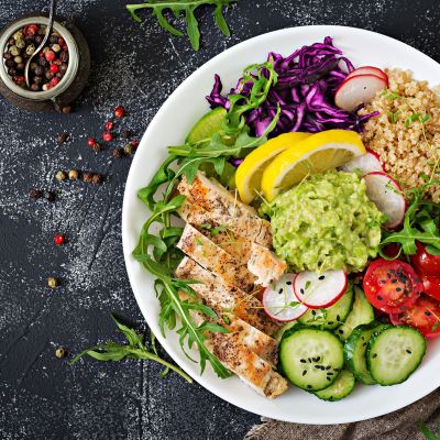 Colorful healthy bowl with grilled chicken, fresh vegetables, quinoa, and guacamole A vibrant plate filled with grilled chicken slices, quinoa, cherry tomatoes, cucumbers with sesame seeds, radishes, shredded purple cabbage, arugula, lime wedges, and a scoop of guacamole arranged beautifully for a nutritious meal.