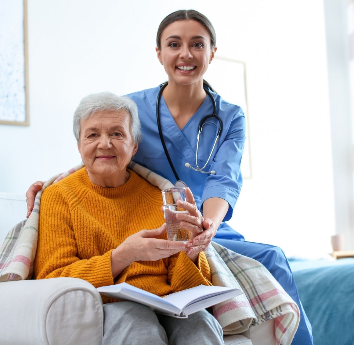 A caregiver in blue scrubs stands behind an elderly woman seated in a cozy chair, gently supporting her as she holds a glass of water. The senior is wrapped in a blanket and reading a book, reflecting comfort, safety, and attentive in-home care.