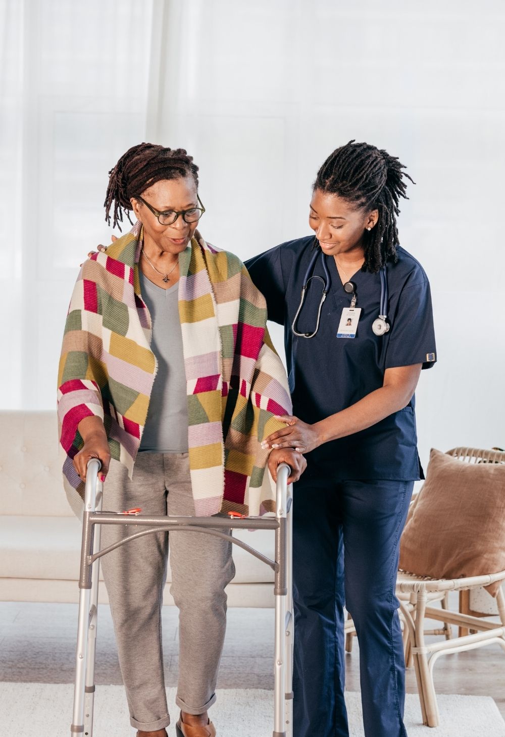 A caregiver in navy scrubs gently supports an elderly woman using a walker. The senior is wrapped in a colorful shawl, and both appear calm and focused, reflecting safe mobility assistance, encouragement, and compassionate in-home care.