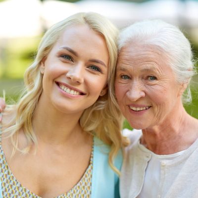 Heartwarming Bond Between Senior and Caregiver Outdoors A smiling young woman stands closely beside an older woman outdoors, both appearing happy and relaxed while enjoying a bright, sunny day.