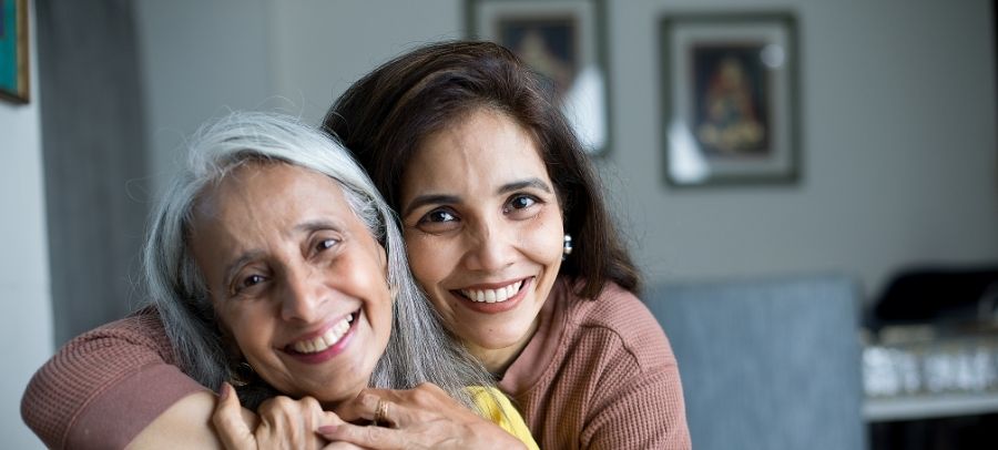 A smiling adult daughter embraces her elderly mother inside their home, showing love, trust, and the comfort of family-centered senior care.