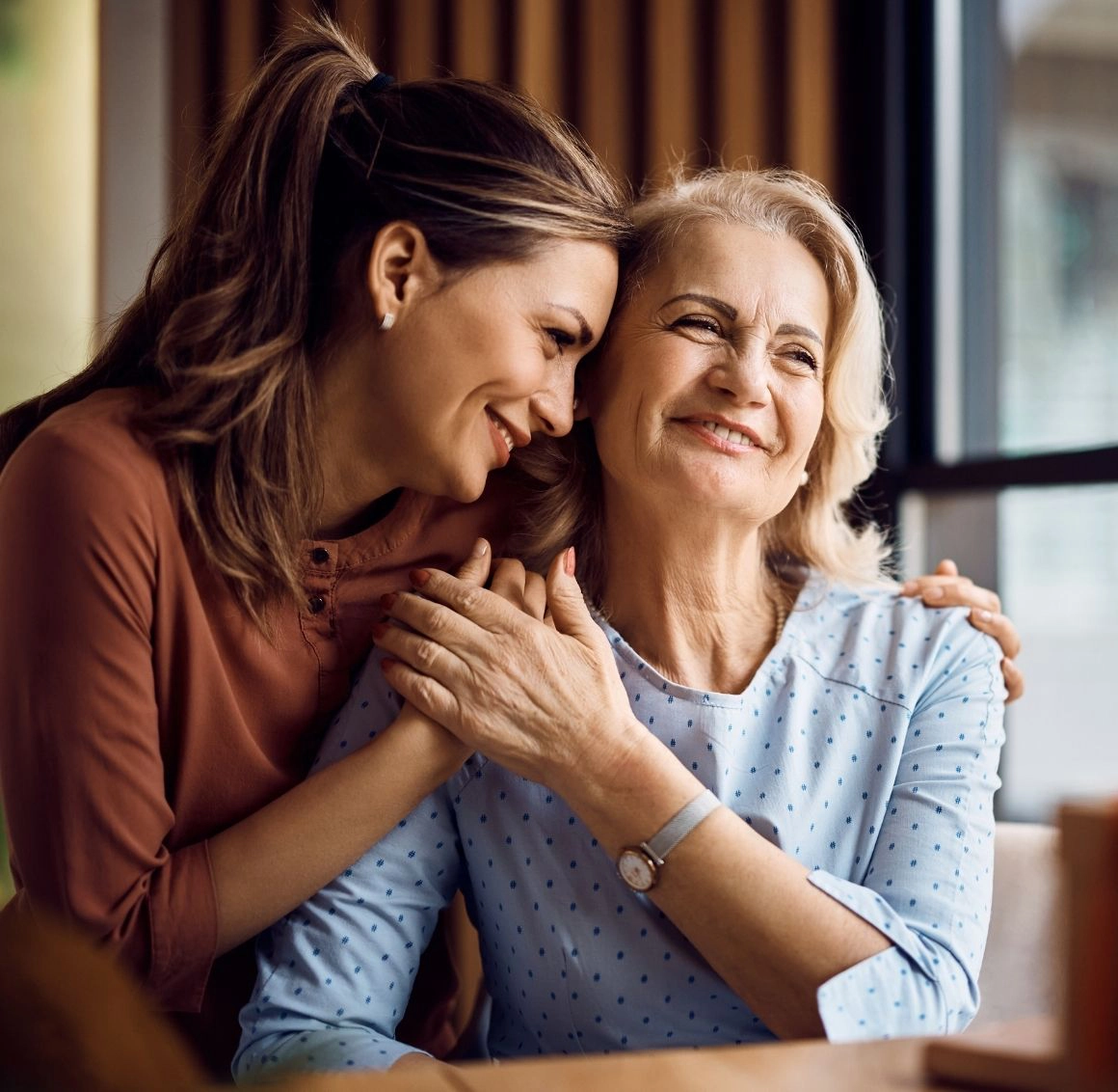 Adult daughter embracing her senior mother with warmth and affection during a heartfelt moment at home An adult daughter gently leans in and holds hands with her smiling elderly mother at a table, showing emotional support, trust, and loving family-centered senior care.