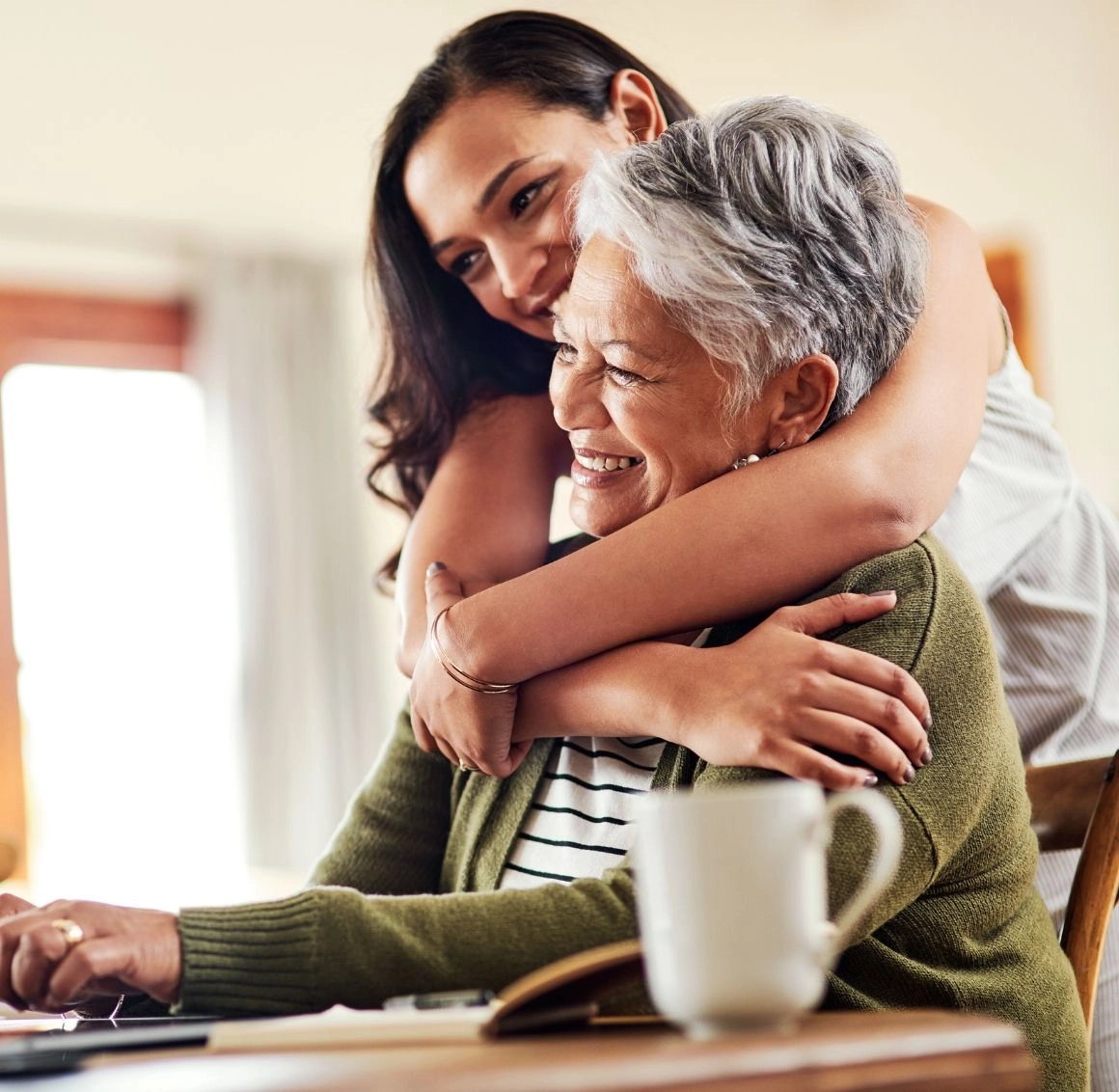 Daughter embracing her senior mother with joy and affection during a warm moment at home A smiling elderly woman sits at a table while her adult daughter hugs her from behind, showing warmth, emotional support, and a strong family connection in senior care.